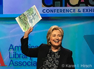 Former Secretary of State Hillary Clinton at the podium raises up her book 'It Takes a Village'